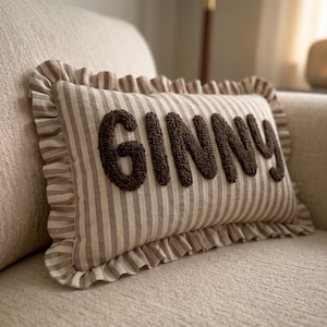 May include: A rectangular throw pillow featuring a beige and brown vertical striped pattern with a ruffled border. The name GINNY is embroidered in dark brown, textured, raised lettering across the center of the pillow fabric surface.