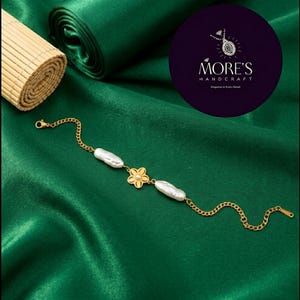 Könnte beinhalten: Ein goldfarbenes Kettenarmband mit zwei länglichen weißen Barockperlen und einem zentralen goldfarbenen Blumenanhänger mit einer kleinen weißen Perle in der Mitte. Das Armband verfügt über einen Karabinerverschluss und eine Verlängerungskette.