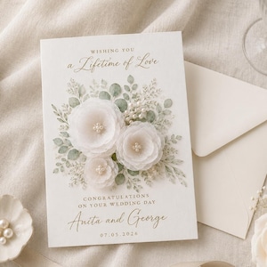 Op de afbeelding: Een witte trouwkaart met een bloemenillustratie van drie witte stoffen bloemen met parelcentra, groene bladeren en de tekst "Wishing You a Lifetime of Love", "CONGRATULATIONS ON YOUR WEDDING DAY", "Anita and George" en "07.05.2026".