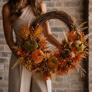 May include: A circular wreath constructed from brown twigs, adorned with velvet pumpkins in shades of olive green and burnt orange, interspersed with artificial autumn maple leaves, dried grasses, small berries, and integrated warm white fairy lights.