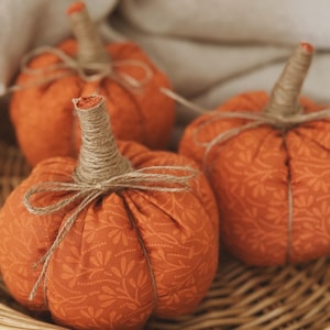 May include: Three fabric pumpkins in a vibrant orange hue, each featuring a subtle tonal floral pattern. The pumpkins are topped with stems wrapped in natural jute twine, which is also tied into a small bow around the base of each stem.