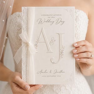 Puede incluir: Una tarjeta de boda color crema con el texto CONGRATULATIONS on your Wedding Day, las iniciales A & J, y los nombres Amelia & Jonathan con la fecha 14TH SEPTEMBER 2026. La tarjeta está decorada con encaje blanco, tul y una perla.