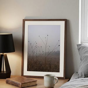 Puede incluir: Una impresión enmarcada de un campo brumoso con plantas altas y delgadas. La obra de arte tiene un marco de madera marrón y un paspartú blanco. Una lámpara negra, un libro antiguo y una taza de cerámica están sobre una mesita de noche de madera.