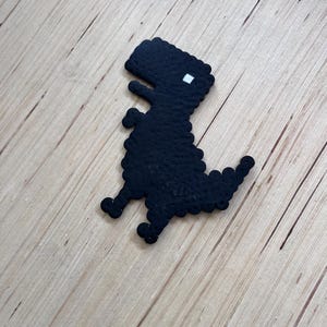 May include: A black pixelated dinosaur figure with a white square eye. The dinosaur is made of small, round beads and is sitting on a light brown wooden surface. The dinosaur has a long tail and small legs.