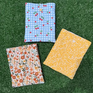 May include: Three rectangular fabric pouches with button closures. One pouch features a light blue and white gingham pattern with red cherries. Another displays a floral print in orange, peach, and green. The third has a yellow floral pattern.