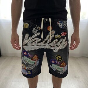 Puede incluir: Pantalones cortos vaqueros negros con m&uacute;ltiples parches y la palabra "Valley" en letras grandes y adornadas. Los pantalones cortos tienen una cintura con cord&oacute;n y varios parches coloridos, incluyendo un s&iacute;mbolo de la paz y una se&ntilde;al de tr&aacute;fico.