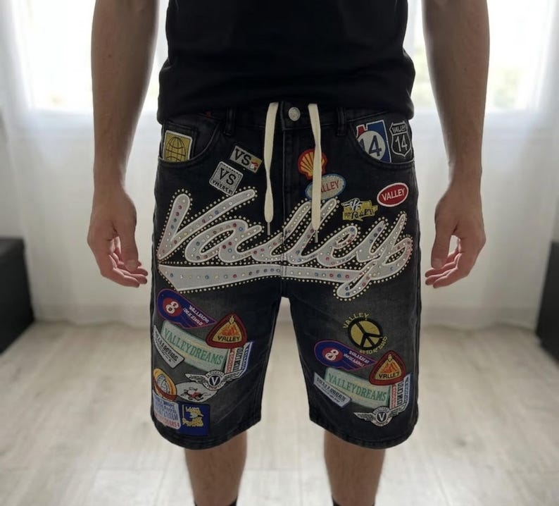 Puede incluir: Pantalones cortos vaqueros negros con m&uacute;ltiples parches y la palabra "Valley" en letras grandes y adornadas. Los pantalones cortos tienen una cintura con cord&oacute;n y varios parches coloridos, incluyendo un s&iacute;mbolo de la paz y una se&ntilde;al de tr&aacute;fico.