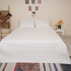 May include: A white quilted bedspread and two matching pillowcases dominate the bedroom. The bed is set against a neutral wall, with a small white side table and decorative items. A wooden ladder and patterned rugs add texture to the scene.
