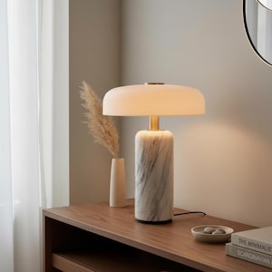 May include: A table lamp featuring a cylindrical white marble base with grey veining, a gold-toned metal neck, and a wide, flat, frosted white glass shade that emits a soft, warm glow, standing on a wooden surface next to a small vase.