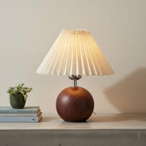 Peut inclure: Une lampe de table dotée d'une base sphérique en bois foncé avec un grain visible et un col chromé. La lampe est surmontée d'un abat-jour en tissu plissé de couleur crème en forme de cône qui diffuse une lueur douce et chaude de l'intérieur.