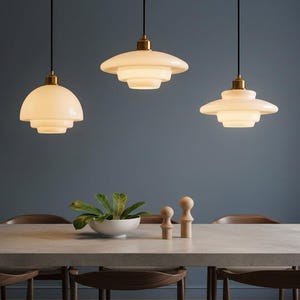 May include: Three pendant lights with tiered, off-white glass shades and brass fittings hang above a dining table. The table is set with a white bowl of greenery and wooden decorative objects. The background is a muted blue.
