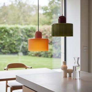 May include: Two contemporary pendant lights suspended from the ceiling. One is orange with a peach-coloured top, the other is olive green with a burgundy top. The lights are cylindrical and hang above a kitchen countertop.