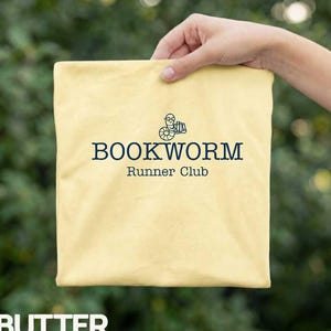 May include: A pale yellow t-shirt featuring a dark blue graphic of a worm wearing glasses and holding a stack of books. Below the graphic, the text reads BOOKWORM Runner Club in a dark blue serif font. The fabric has a smooth, matte finish.