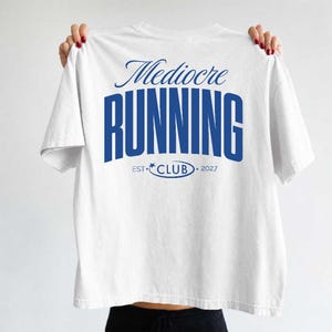May include: A white short-sleeved t-shirt featuring blue text on the back. The graphic displays the words Mediocre RUNNING in a bold, stacked font, with the text EST 2027 and the word CLUB enclosed in an oval with a small star icon.