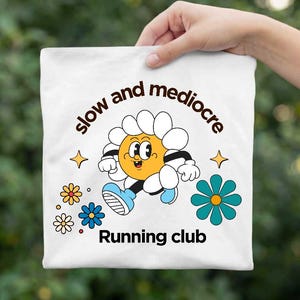 May include: A white t-shirt featuring a graphic of a cartoon daisy with a yellow face and white petals, wearing blue sneakers. The text reads "slow and mediocre" in an arc above and "Running club" below, surrounded by small colorful flowers.