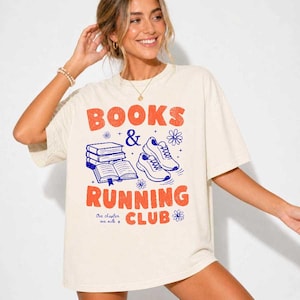 May include: An off-white short-sleeve t-shirt featuring a graphic print in orange and blue. The design includes the text "BOOKS & RUNNING CLUB" with "One chapter one mile" below, alongside illustrations of a stack of books and two running shoes.
