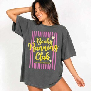May include: A charcoal gray short-sleeve t-shirt featuring a graphic design with vertical pink stripes, yellow cursive text that reads Books Running Club, and the text ONE CHAPTER ONE MILE in yellow, accented with small white stars.