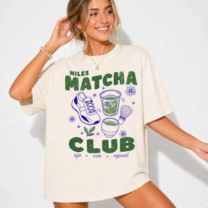 May include: An off-white short-sleeve t-shirt featuring a graphic print with the text MILES MATCHA CLUB and sip + run + repeat. The design includes a running shoe, a glass of matcha, a matcha whisk, a small bowl, and floral accents in green and blue.