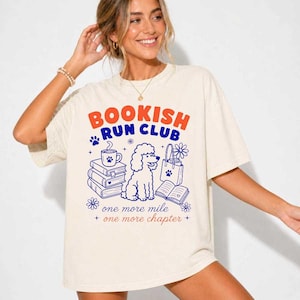 May include: An off-white short-sleeve t-shirt featuring a graphic print with the text "BOOKISH RUN CLUB" in orange and blue. The design includes a poodle, a stack of books, a coffee mug, a tote bag, and the text "one more mile one more chapter."