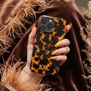 May include: A rectangular smartphone case featuring a brown and black tortoiseshell pattern with the gold-colored letters KKC printed near the bottom. The case has a cutout for a triple-lens camera system on the upper left corner of the back panel.