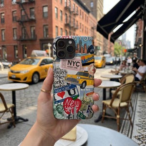 May include: A smartphone case featuring a collage of New York City imagery, including a yellow taxi, the Statue of Liberty, a red heart icon, and text elements like NYC, Metro, and New York, all set against a multi-colored urban graphic background.
