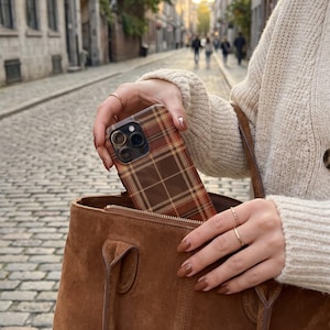 Puede incluir: Una funda para smartphone con un patr&oacute;n de cuadros marr&oacute;n, naranja y amarillo, que se introduce en un bolso de ante marr&oacute;n. La funda tiene un recorte para un sistema de triple c&aacute;mara y est&aacute; sujeta por dedos con esmalte marr&oacute;n.