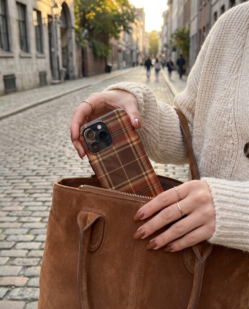 Puede incluir: Una funda para smartphone con un patr&oacute;n de cuadros marr&oacute;n, naranja y amarillo, que se introduce en un bolso de ante marr&oacute;n. La funda tiene un recorte para un sistema de triple c&aacute;mara y est&aacute; sujeta por dedos con esmalte marr&oacute;n.