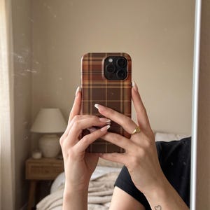 Puede incluir: Una funda para smartphone con un patr&oacute;n de cuadros marr&oacute;n y beige y un recorte para el m&oacute;dulo de c&aacute;mara en marr&oacute;n oscuro. La funda se muestra exhibiendo un dise&ntilde;o de tart&aacute;n cl&aacute;sico con l&iacute;neas entrecruzadas en tonos marrones, beige y naranja.