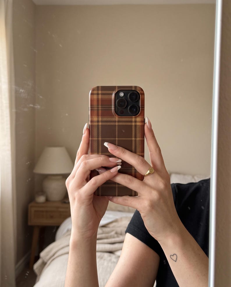 Puede incluir: Una funda para smartphone con un patr&oacute;n de cuadros marr&oacute;n y beige y un recorte para el m&oacute;dulo de c&aacute;mara en marr&oacute;n oscuro. La funda se muestra exhibiendo un dise&ntilde;o de tart&aacute;n cl&aacute;sico con l&iacute;neas entrecruzadas en tonos marrones, beige y naranja.