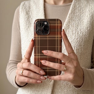 Puede incluir: Una funda para smartphone con un patr&oacute;n de cuadros marrones y tostados con una cuadr&iacute;cula de l&iacute;neas que se cruzan. La funda tiene un recorte para un sistema de c&aacute;mara de triple lente en la parte posterior. El dise&ntilde;o cubre toda la superficie.