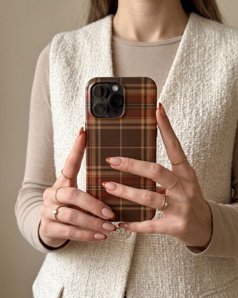 Puede incluir: Una funda para smartphone con un patr&oacute;n de cuadros marrones y tostados con una cuadr&iacute;cula de l&iacute;neas que se cruzan. La funda tiene un recorte para un sistema de c&aacute;mara de triple lente en la parte posterior. El dise&ntilde;o cubre toda la superficie.