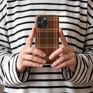 Puede incluir: Una funda para smartphone con un patr&oacute;n de cuadros en tonos marr&oacute;n, beige y naranja. La funda tiene forma rectangular con esquinas redondeadas y un recorte para el conjunto de lentes de la c&aacute;mara en la parte superior izquierda.