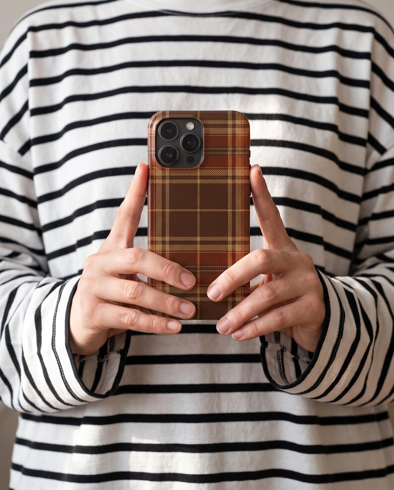 Puede incluir: Una funda para smartphone con un patr&oacute;n de cuadros en tonos marr&oacute;n, beige y naranja. La funda tiene forma rectangular con esquinas redondeadas y un recorte para el conjunto de lentes de la c&aacute;mara en la parte superior izquierda.