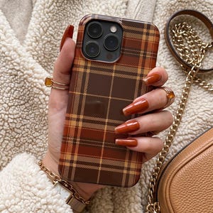 Puede incluir: Una funda para smartphone con un patr&oacute;n de cuadros en tonos marr&oacute;n, naranja y beige. La funda tiene forma rectangular con esquinas redondeadas y un recorte para un sistema de triple c&aacute;mara en la parte posterior. El dise&ntilde;o consta de l&iacute;neas.