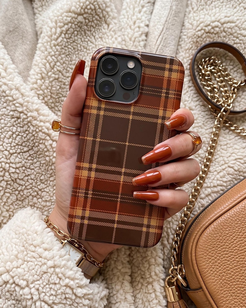 Puede incluir: Una funda para smartphone con un patr&oacute;n de cuadros en tonos marr&oacute;n, naranja y beige. La funda tiene forma rectangular con esquinas redondeadas y un recorte para un sistema de triple c&aacute;mara en la parte posterior. El dise&ntilde;o consta de l&iacute;neas.