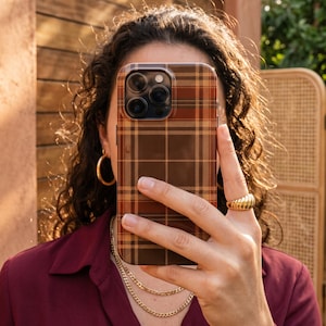 Puede incluir: Una funda rectangular para smartphone con un patr&oacute;n de cuadros en tonos marr&oacute;n, beige y naranja. La funda tiene un recorte para un sistema de triple c&aacute;mara en la esquina superior izquierda y cubre la parte trasera del dispositivo con un acabado liso.