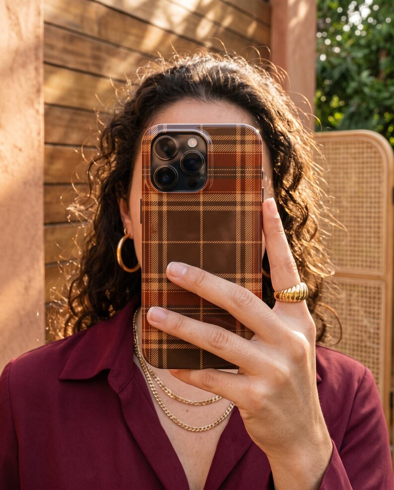 Puede incluir: Una funda rectangular para smartphone con un patr&oacute;n de cuadros en tonos marr&oacute;n, beige y naranja. La funda tiene un recorte para un sistema de triple c&aacute;mara en la esquina superior izquierda y cubre la parte trasera del dispositivo con un acabado liso.