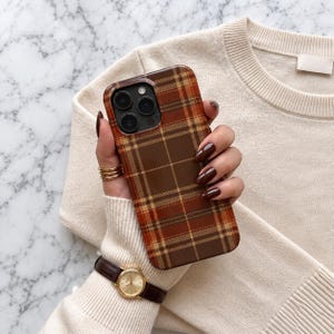 May include: A smartphone case featuring a brown, orange, and tan plaid pattern. The case has a cutout for a triple-lens camera system on the back. The design consists of intersecting horizontal and vertical lines forming a classic tartan grid.