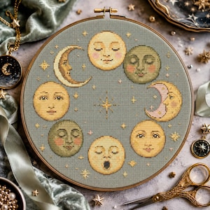 May include: A cross-stitch embroidery hoop with a celestial design. The artwork features various moon phases with smiling faces, set against a muted teal background with star accents. The wooden hoop is approximately 20 cm in diameter.