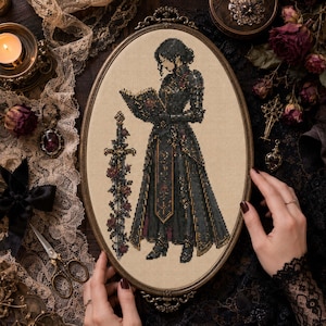 May include: An oval framed cross-stitch depicting a figure in a long black gown reading a book, with a sword adorned with roses. The frame is held by hands with dark nail polish. The background includes lace, a candle, and other decorative items.