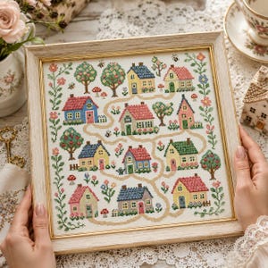 May include: A framed cross-stitch featuring a village scene with colorful houses, trees, and a winding path. The houses have red, blue, yellow, and pink roofs. The frame is a light wood color, and the artwork is surrounded by floral and leaf patterns.