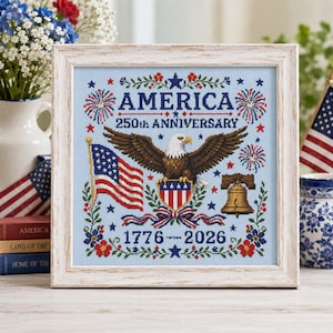 May include: Framed cross-stitch artwork celebrating America's 250th Anniversary. The design features an American flag, an eagle, the Liberty Bell, and the years 1776-2026. The word "AMERICA" is prominently displayed, surrounded by fireworks and floral elements.