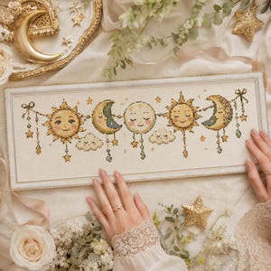 May include: A framed cross-stitch artwork depicting smiling suns, moons, and clouds, with dangling stars and beads. The design uses soft yellow, cream, and blue colours. The artwork is presented on a cream-coloured fabric.