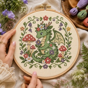 Peut inclure: Un cercle de broderie au point de croix avec un dragon vert orné de fleurs et d'ailes, entouré de champignons à chapeaux rouges et de motifs floraux. Le motif est sur un tissu crème, maintenu dans un cercle en bois.