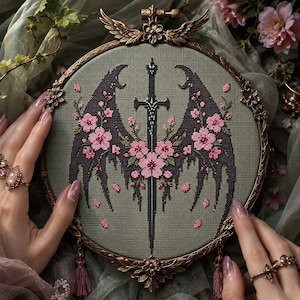 May include: Cross-stitch embroidery featuring a black sword and stylized wings adorned with pink cherry blossoms. The design is set in a round, ornate gold frame with decorative elements and tassels.