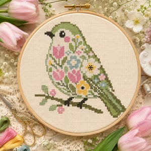 May include: A cross-stitch embroidery of a bird perched on a branch, set in a wooden hoop. The bird is green with a floral design of pink, yellow, and blue flowers. The background is a light beige fabric.