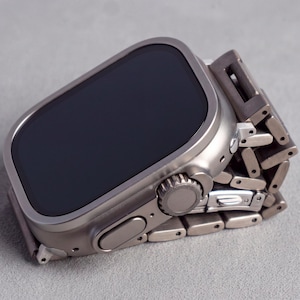 Könnte beinhalten: Eine titanfarbene Smartwatch mit einem rechteckigen schwarzen Display und einem passenden Metallgliederarmband. Das Gerät verfügt über eine markante seitliche Krone, eine flache Taste und ein robustes Gehäuse mit sichtbaren Schrauben.