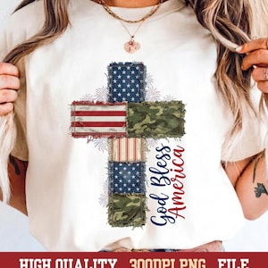 May include: A cream-colored t-shirt featuring a central cross graphic composed of distressed fabric patches in American flag patterns and green camouflage. The text God Bless America is printed in blue and red script next to the cross.