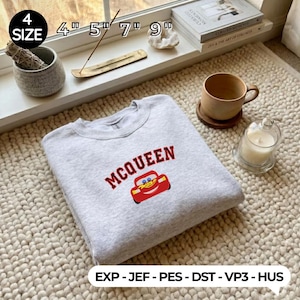 Puede incluir: Una sudadera gris claro con cuello redondo con la palabra "MCQUEEN" en letras granates y un gráfico de coche de dibujos animados rojo. La sudadera se muestra sobre una alfombra texturizada, con una vela, una taza y libros en el fondo.