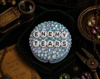 Op de afbeelding: Een ronde telefoongreep bedekt met iriserende blauwe strass-steentjes met witte plastic letterkralen die de woorden ENEMY TEARS in twee rijen spellen. Het accessoire heeft een reflecterend, veelkleurig oppervlak dat het licht opvangt over de textuur van de bovenkant.